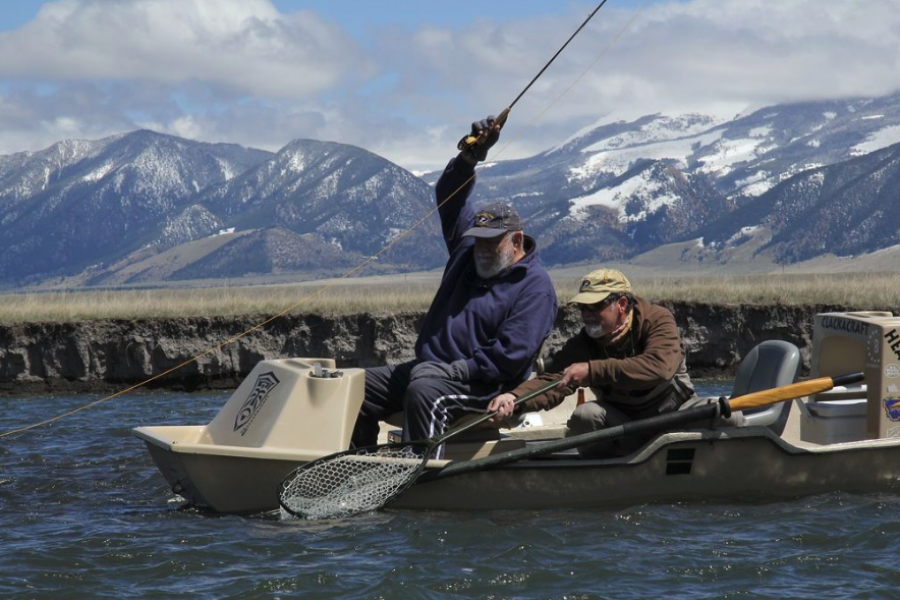 Man in a drift boat with arm in the air fishing fish while guide nets the fish in the water on fly fishing tour in montana with snow capped mountains in the background.